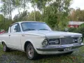 Vit Plymouth Barracuda från 1960-talet parkerad på en gräsmatta med träd och en röd byggnad i bakgrunden.