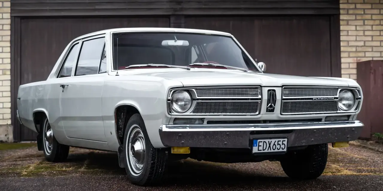 En vit, klassisk Plymouth Valiant står parkerad framför ett garage.