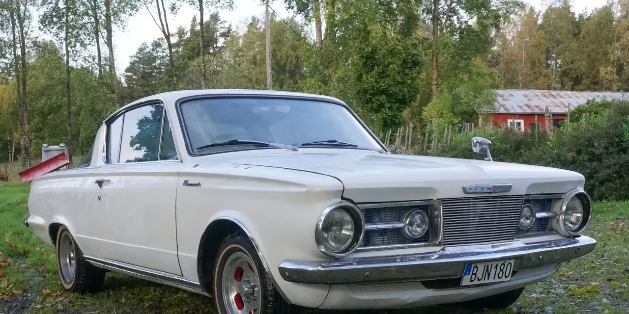 Vit Plymouth Barracuda från 1960-talet parkerad på en gräsmatta med träd och en röd byggnad i bakgrunden.