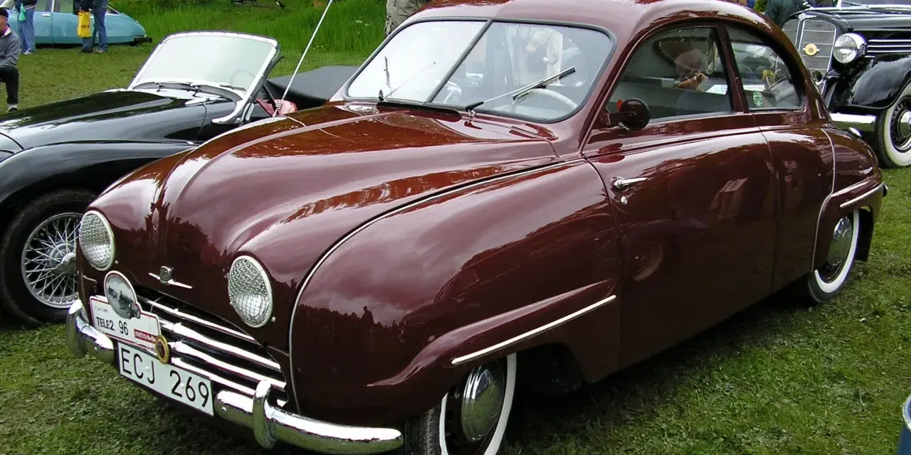 En vinröd Saab 92 står på gräs, med en svart cabriolet bredvid. Bilen har en klassisk design och en svensk registreringsskylt.