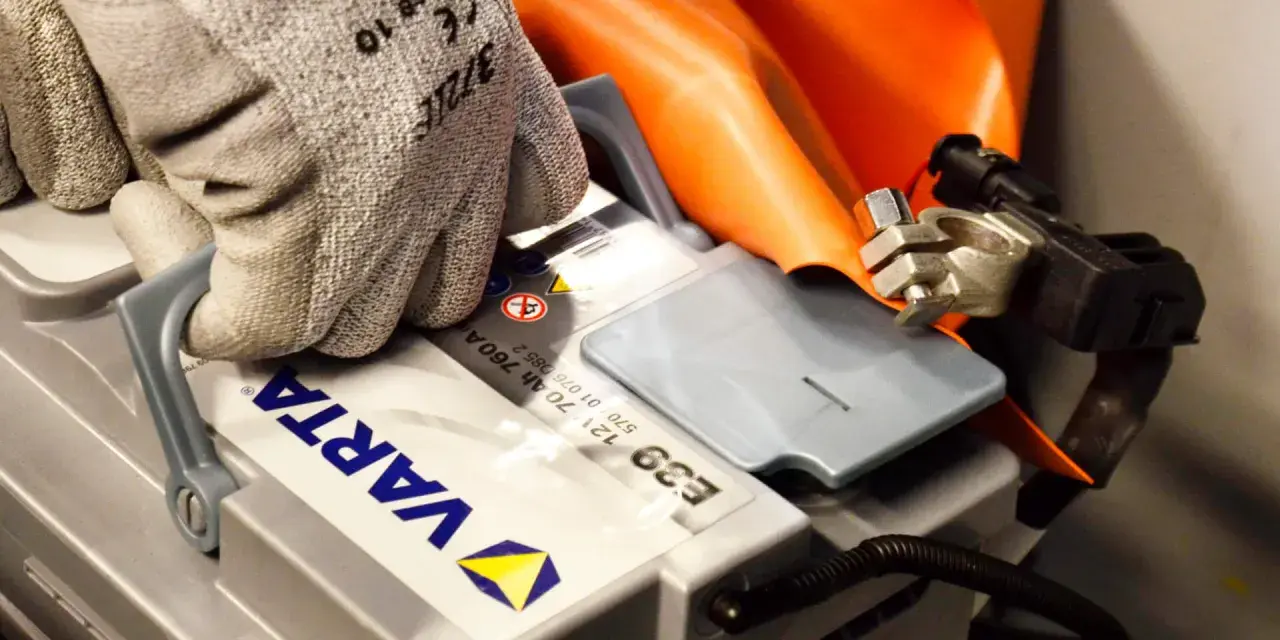 Hand med skyddshandske kopplar ihop kablar för att ladda bilbatteri. Orange skyddsdyna syns bakom.