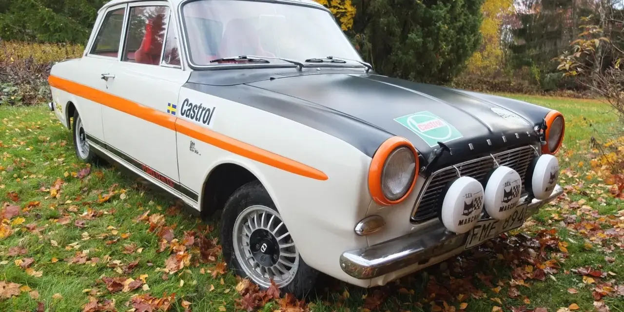 Ford Taunus 12M i rallystuk, med orange detaljer och Castrol-logga, står på en gräsmatta täckt av höstlöv.