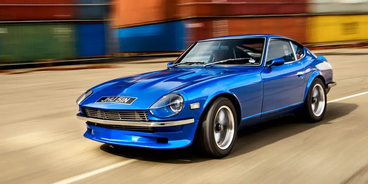 Blå Nissan Datsun 240Z kör förbi färgglada fraktcontainrar.