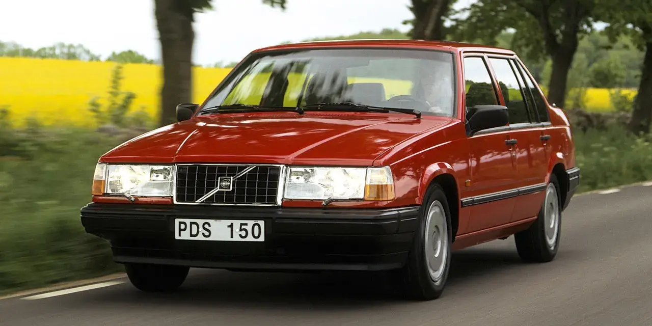 En röd 740 Volvo kör längs en landsväg, med en gul rapsåker och träd i bakgrunden.