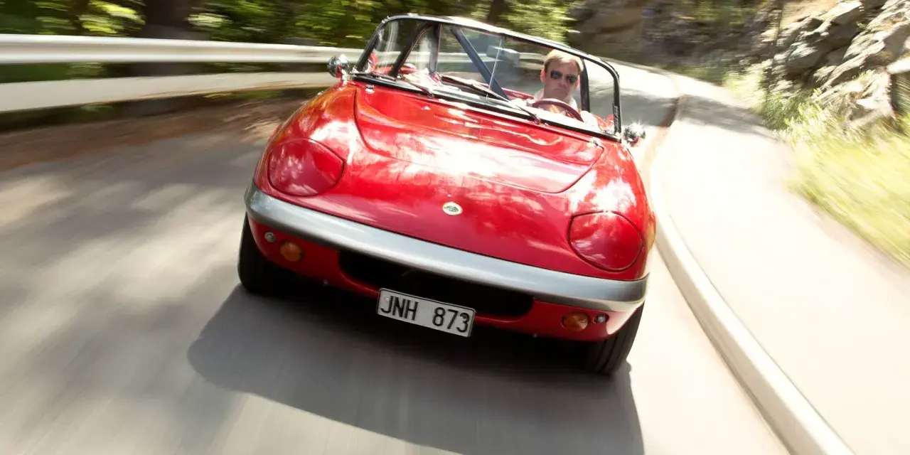 Röd Lotus Elan M100 körs på en kurvig väg genom en skog. Föraren bär solglasögon.
