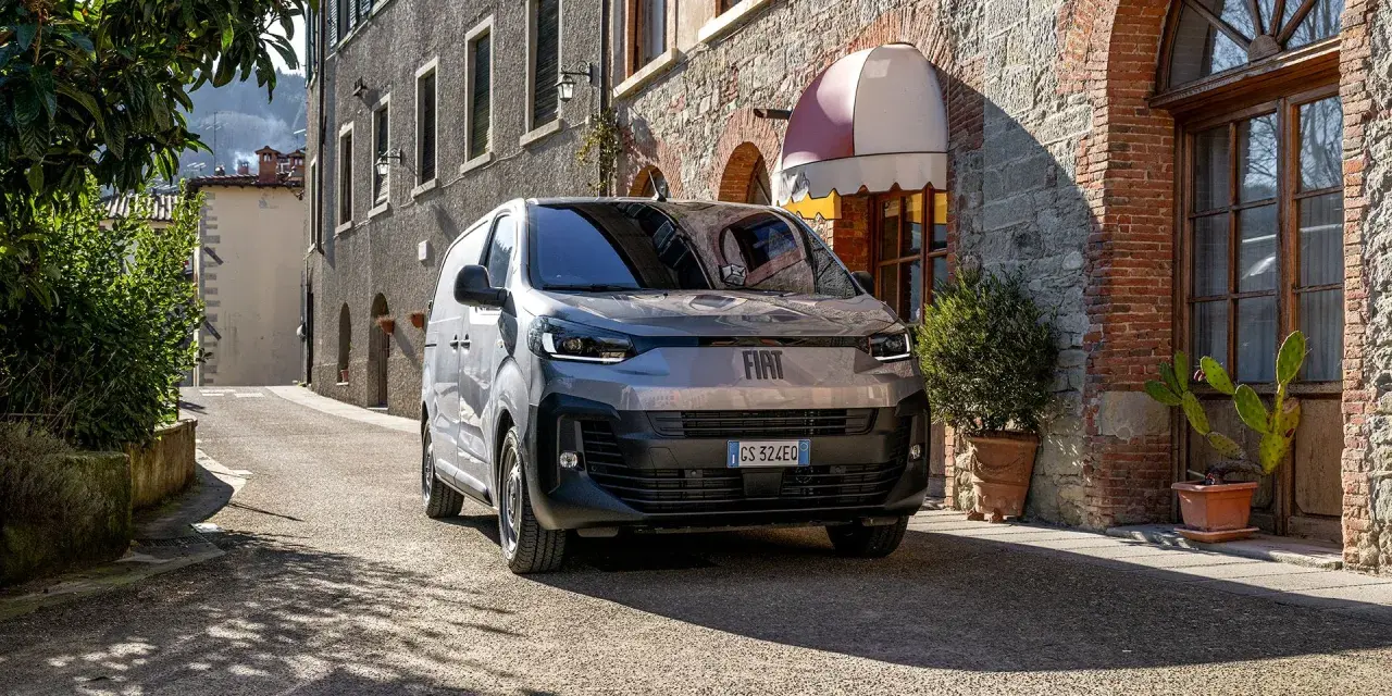 En grå Fiat Doblo står parkerad på en smal gata i Italien. En vacker byggnad med tegel och sten syns i bakgrunden.