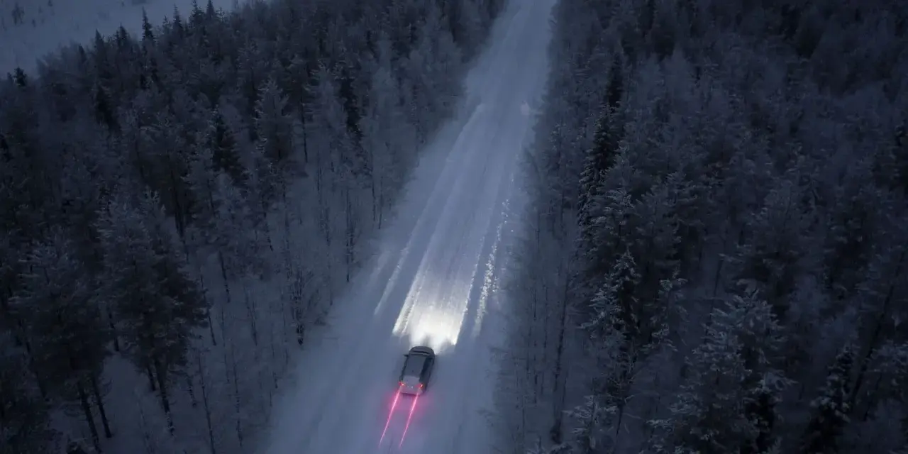 Bil kör på snöig skogsväg i mörker, halvljus symbolen lyser upp vägen.