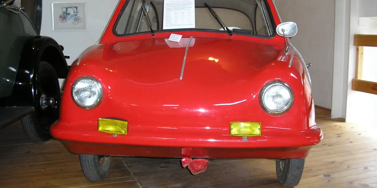 En röd, rundad bil, en Messerschmitt KR200, står på ett trägolv. Fram king Fulda.