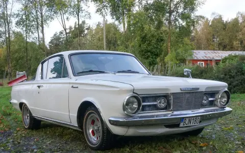 Vit Plymouth Barracuda från 1960-talet parkerad på en gräsmatta med träd och en röd byggnad i bakgrunden.