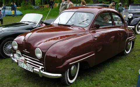 En vinröd Saab 92 står på gräs, med en svart cabriolet bredvid. Bilen har en klassisk design och en svensk registreringsskylt.
