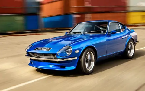 Blå Nissan Datsun 240Z kör förbi färgglada fraktcontainrar.