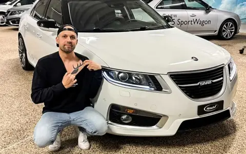 Man med hjort-tatuering på bröstet poserar framför en vit Saab.