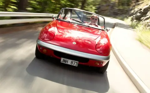 Röd Lotus Elan M100 körs på en kurvig väg genom en skog. Föraren bär solglasögon.