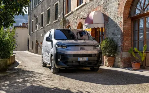 En grå Fiat Doblo står parkerad på en smal gata i Italien. En vacker byggnad med tegel och sten syns i bakgrunden.