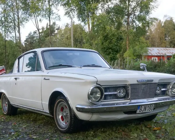 Vit Plymouth Barracuda från 1960-talet parkerad på en gräsmatta med träd och en röd byggnad i bakgrunden.
