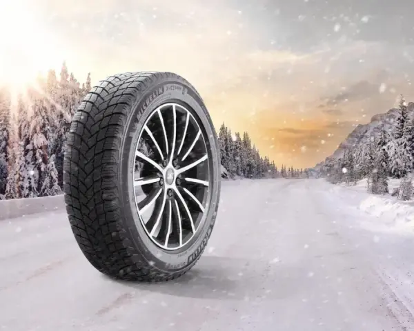 Michelin X-Ice Snow-däck på en snötäckt väg i en vinterskog. Solen bryter igenom molnen.