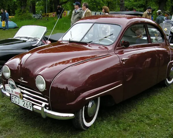En vinröd Saab 92 står på gräs, med en svart cabriolet bredvid. Bilen har en klassisk design och en svensk registreringsskylt.