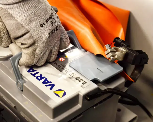 Hand med skyddshandske kopplar ihop kablar för att ladda bilbatteri. Orange skyddsdyna syns bakom.