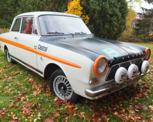 Ford Taunus 12M i rallystuk, med orange detaljer och Castrol-logga, står på en gräsmatta täckt av höstlöv.