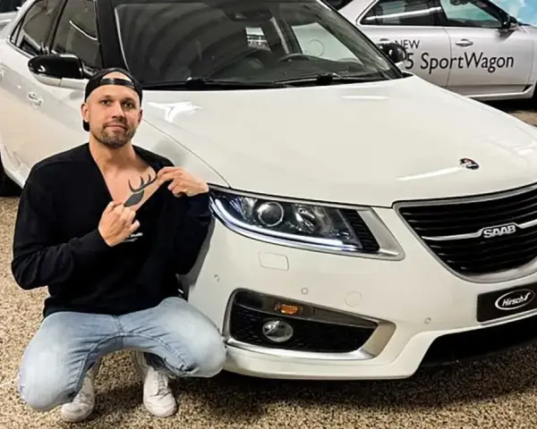Man med hjort-tatuering på bröstet poserar framför en vit Saab.