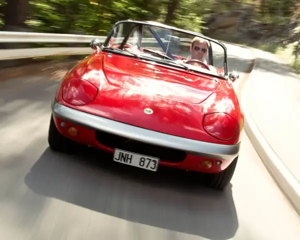 Röd Lotus Elan M100 körs på en kurvig väg genom en skog. Föraren bär solglasögon.