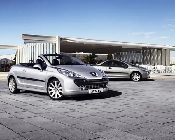 Två silverfärgade Peugeot 207 CC cabriolet står parkerade på en stenlagd uteplats framför en modern byggnad.
