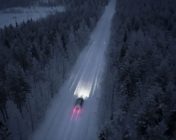 Bil kör på snöig skogsväg i mörker, halvljus symbolen lyser upp vägen.