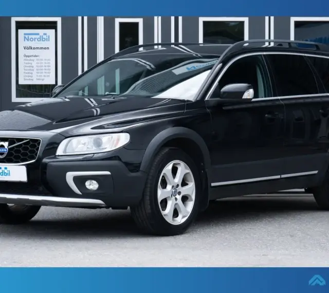 Svart Volvo XC70 parkerad framför en byggnad med skylten "Nordbil Välkommen". Bilen har takräcken och en robust design.