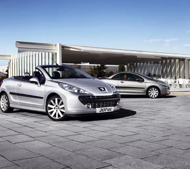 Två silverfärgade Peugeot 207 CC cabriolet står parkerade på en stenlagd uteplats framför en modern byggnad.