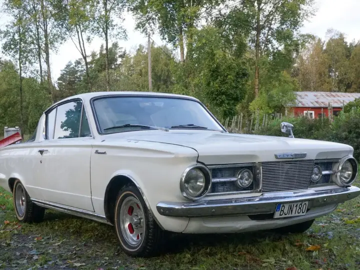 Vit Plymouth Barracuda från 1960-talet parkerad på en gräsmatta med träd och en röd byggnad i bakgrunden.