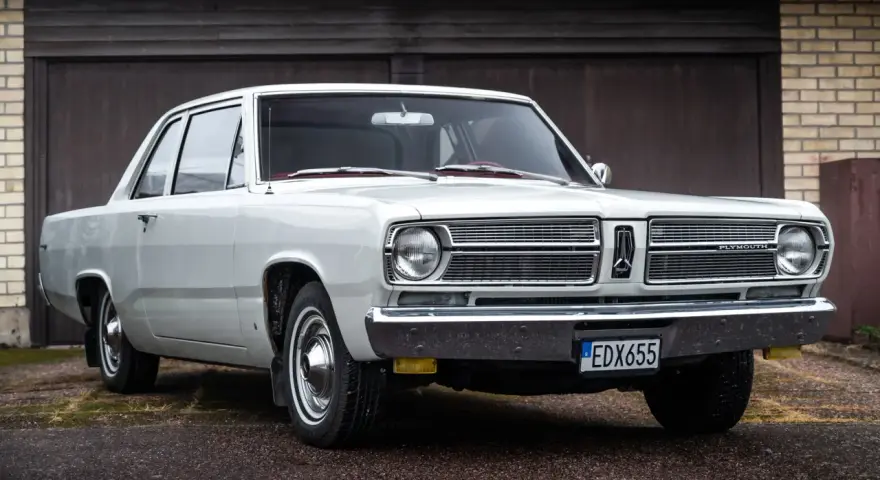En vit, klassisk Plymouth Valiant står parkerad framför ett garage.
