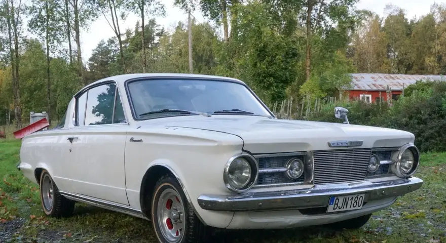 Vit Plymouth Barracuda från 1960-talet parkerad på en gräsmatta med träd och en röd byggnad i bakgrunden.