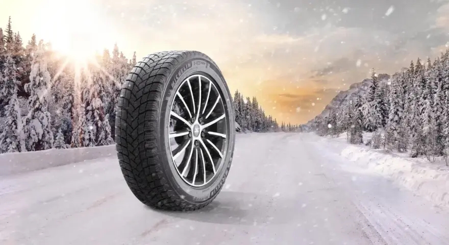 Michelin X-Ice Snow-däck på en snötäckt väg i en vinterskog. Solen bryter igenom molnen.