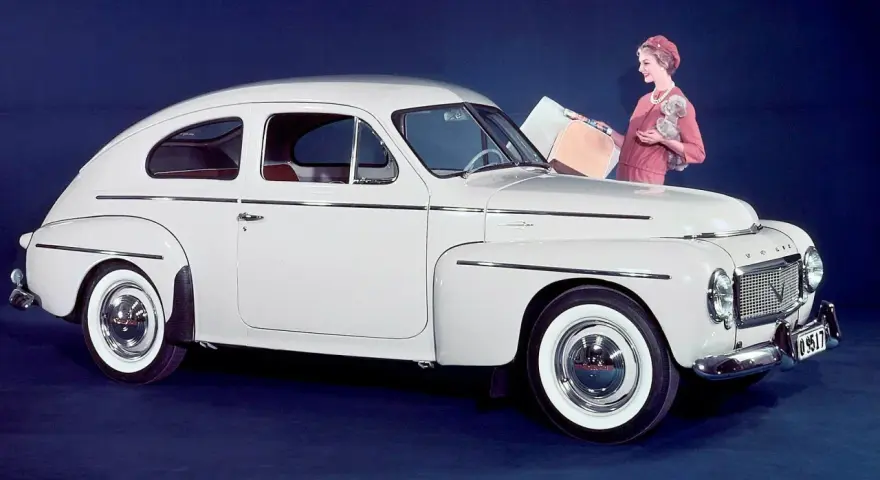 En elegant vit Volvo PV 444 med en kvinna och en hund bredvid.