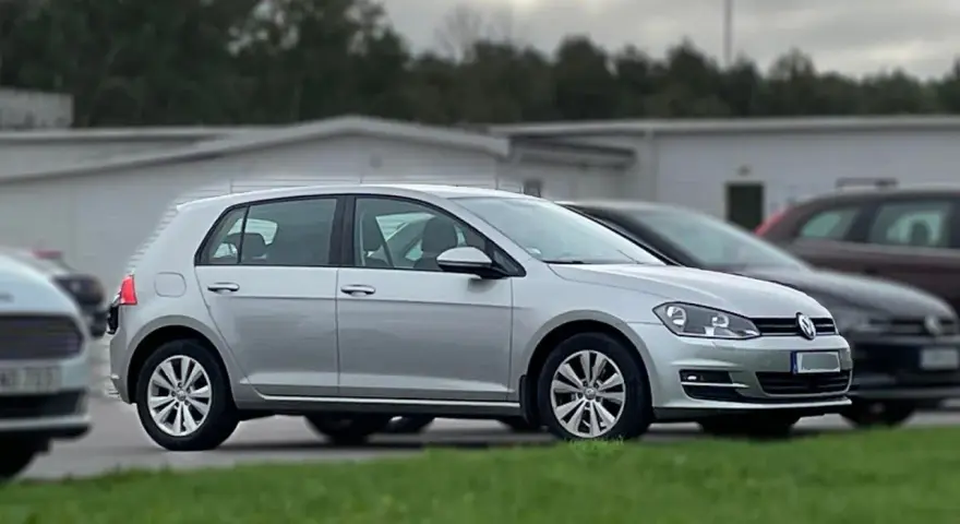 Silver Golf sportsvan parkerad på gräs. En mörkare bil syns bakom.