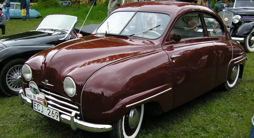 En vinröd Saab 92 står på gräs, med en svart cabriolet bredvid. Bilen har en klassisk design och en svensk registreringsskylt.