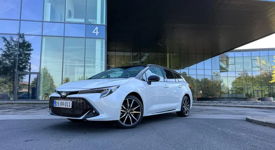 Vit Toyota Corolla Touring Sports framför en modern glasbyggnad. Inga problem med denna bil, bara ren stil.