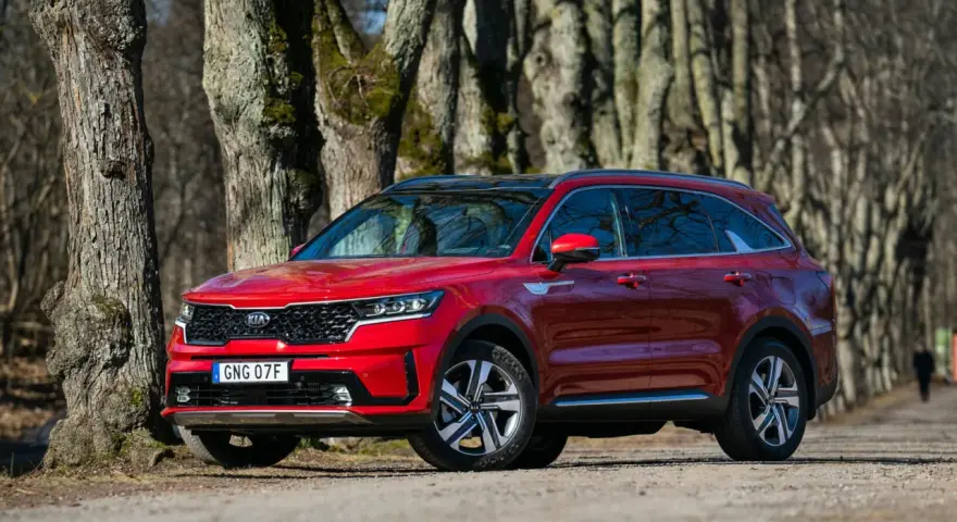 Röd Kia Sorento parkerad längs en trädkantad grusväg. En person syns i bakgrunden, kanske på väg att undersöka ett eventuellt kia sorento problem.