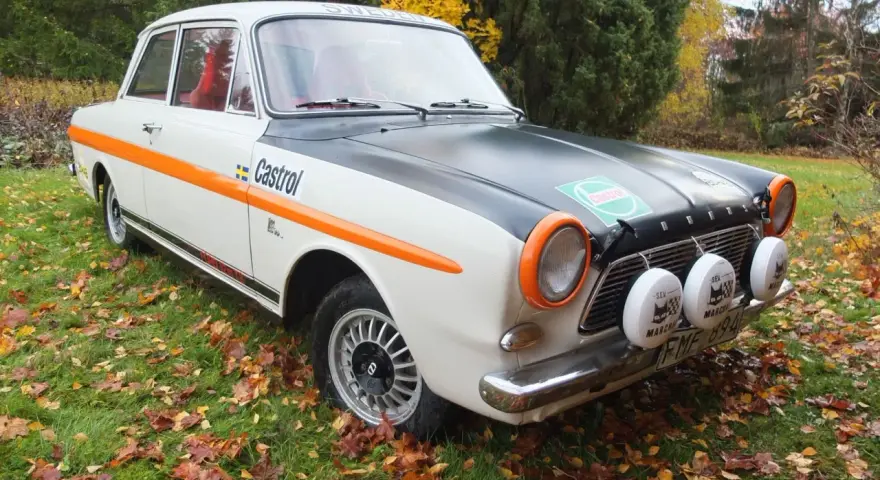 Ford Taunus 12M i rallystuk, med orange detaljer och Castrol-logga, står på en gräsmatta täckt av höstlöv.