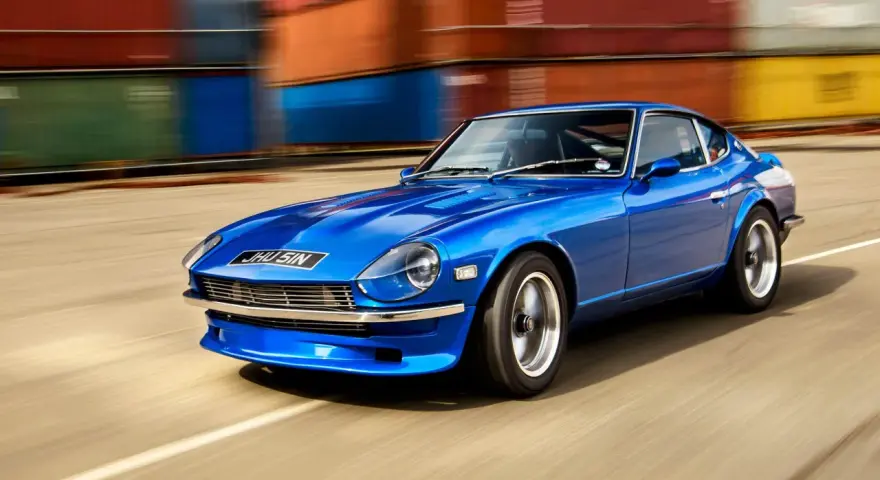 Blå Nissan Datsun 240Z kör förbi färgglada fraktcontainrar.
