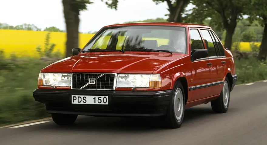 En röd 740 Volvo kör längs en landsväg, med en gul rapsåker och träd i bakgrunden.