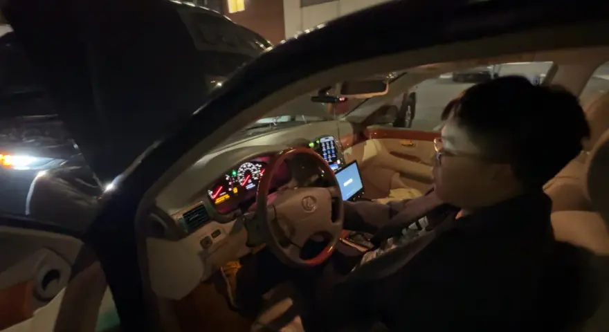 Personen försöker starta bilen, men den startar inte, bara klickar. Motorhuven är öppen och en laptop är på passagerarsätet.