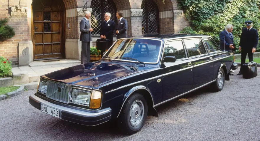 Mörkblå Volvo 264 Bertone limousine parkerad framför en byggnad med män i kostym.