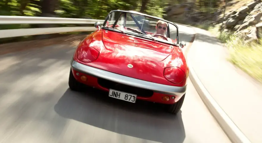 Röd Lotus Elan M100 körs på en kurvig väg genom en skog. Föraren bär solglasögon.