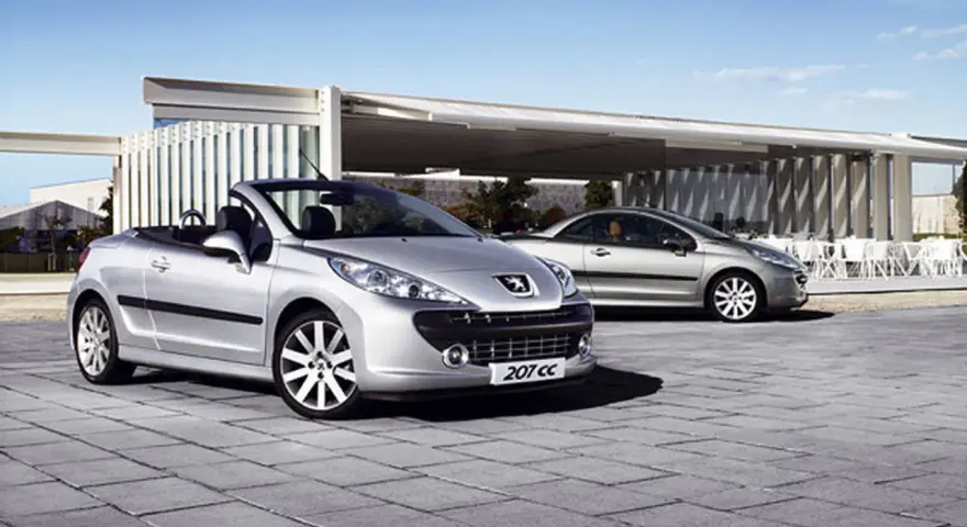 Två silverfärgade Peugeot 207 CC cabriolet står parkerade på en stenlagd uteplats framför en modern byggnad.