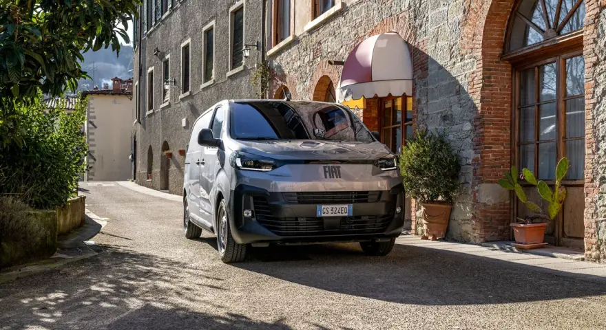 En grå Fiat Doblo står parkerad på en smal gata i Italien. En vacker byggnad med tegel och sten syns i bakgrunden.