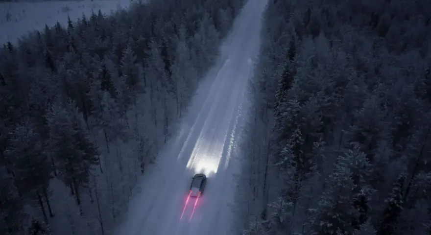 Bil kör på snöig skogsväg i mörker, halvljus symbolen lyser upp vägen.