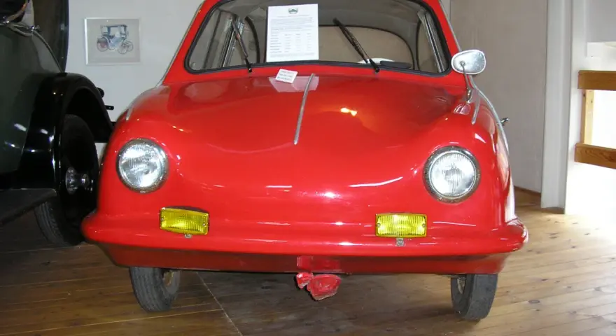 En röd, rundad bil, en Messerschmitt KR200, står på ett trägolv. Fram king Fulda.