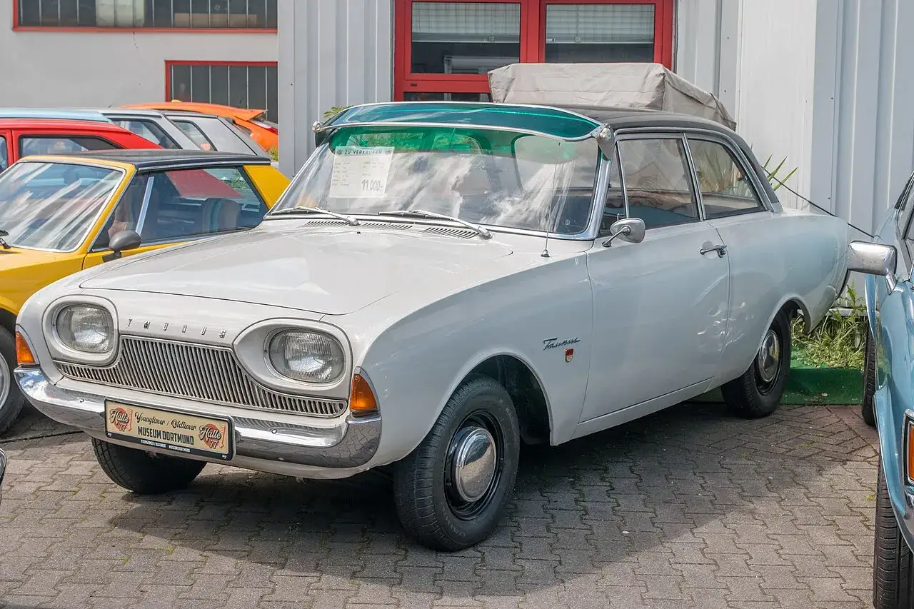 Vit Ford Taunus 1960 med gr&ouml;n solsk&auml;rm, parkerad bredvid andra klassiska bilar.