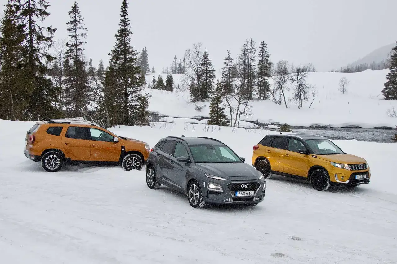 Tre bilar, en orange Dacia Duster, en gr&aring; Hyundai Kona och en gul Suzuki Vitara, st&aring;r parkerade i sn&ouml;n under ett vintertest.